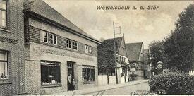 1935 Wewelsfleth; Dorfstraße mit im Vordergrund dem Geschäfthaus „H. Kröger Söhne – Kolonial- und Eisenwaren“