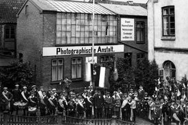 1923 Vor der Photographischen Anstalt von Ludwig Behning am Markt ist die Musikgruppe (Trommler und Pfeifer Corps) der Kinder-Schützengilde mit Flaggen, Standarten und dem hölzernen Adler sowie einer aus Nadelgehölz gebundenen Krone angetreten